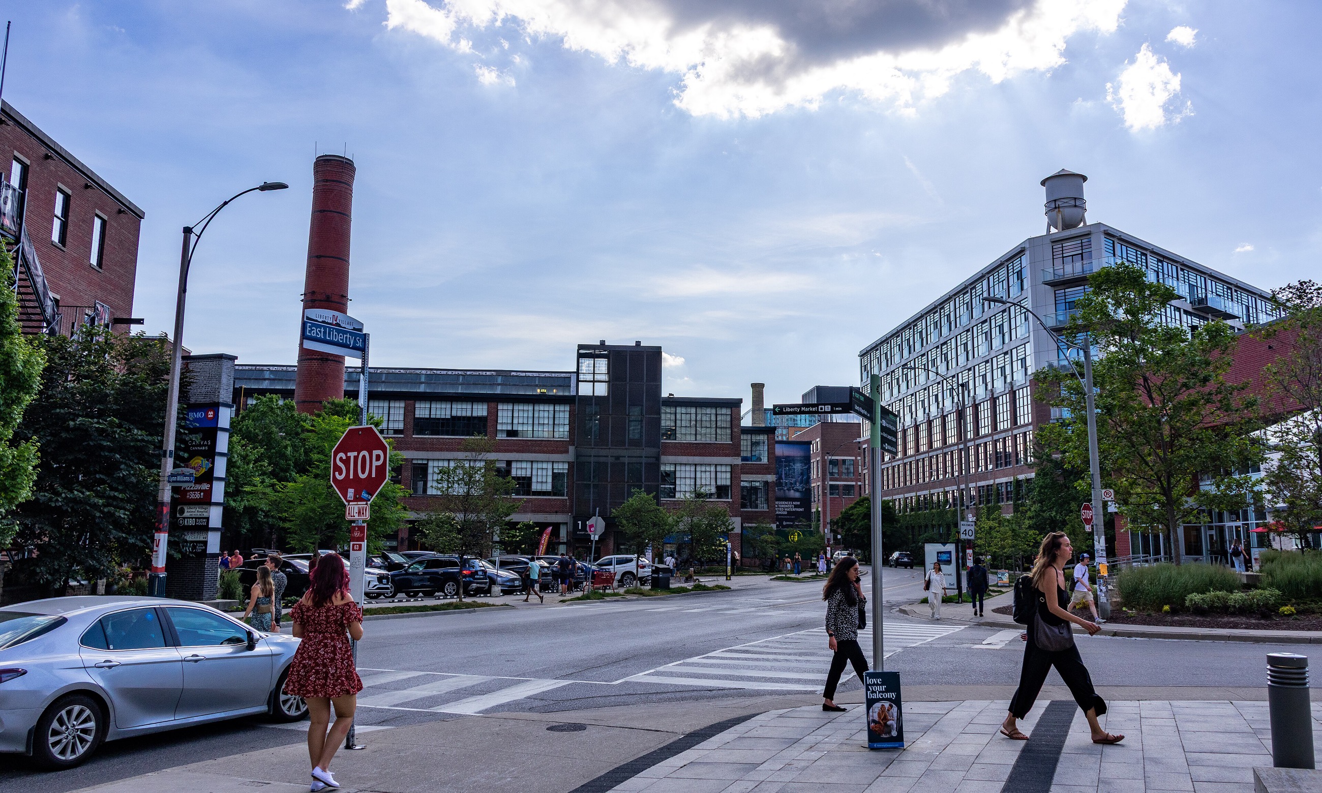 The East Liberty Street corner in Liberty Village on a summer afternoon, with pedestrians crossing between converted brick factory buildings, a smokestack and rooftop water tower in view